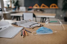 foto van een mondkapje een,bril, potloden, pennen en 2 open geslagen boeken op een tafel in een klaslokaal
