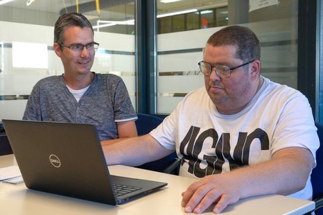 2 mannen zitten achter een bureau en kijken naar een laptop